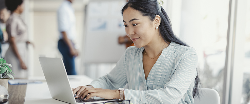 Asian woman working on laptop. Businesswoman busy working on laptop computer at office with colleagues in the background.